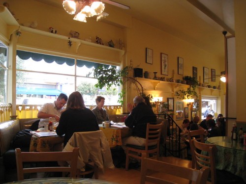 A cozy café interior with yellow walls, tables occupied by patrons, and plants decorating the windowsill.