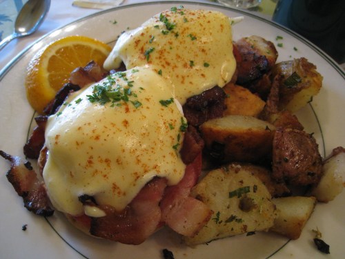 A plate of eggs Benedict with ham, hollandaise sauce, and seasoned potatoes, garnished with parsley and an orange slice.