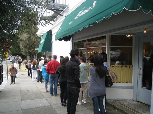 A long line of people waiting outside a restaurant with green awnings on a city street.