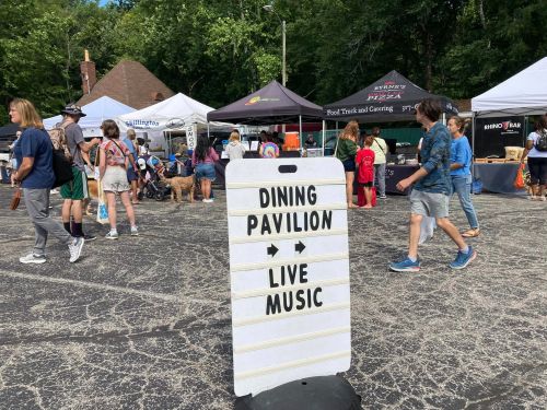 A sign points to a dining pavilion and live music at a bustling outdoor market with people and tents.