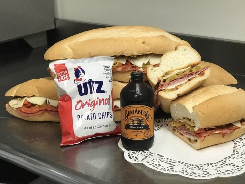 A stack of large sandwiches with meats and cheese, a bag of Utz potato chips, and a bottle of Bundaberg root beer.