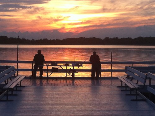 Two people fishing on a dock at sunset, with a calm lake reflecting the colorful sky. Benches line the wooden platform.