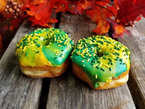Two donuts with green and yellow icing, topped with green and yellow sprinkles, on a wooden surface with autumn leaves.