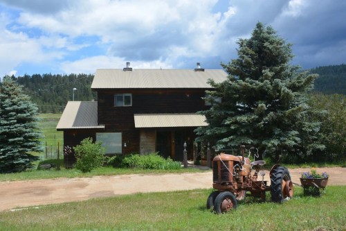 A rustic wooden house with a metal roof, surrounded by trees and a vintage tractor in the foreground.