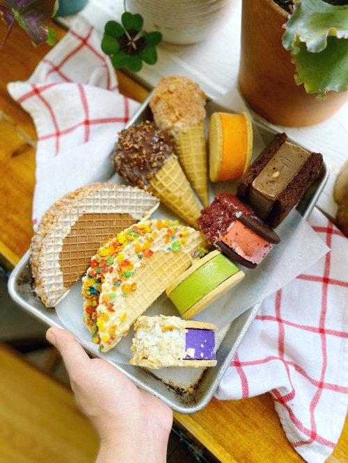 A hand holding a tray of colorful desserts, including ice cream cones and various cake slices, on a checkered cloth.
