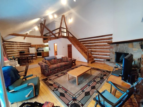 Cozy cabin interior with wooden beams, a staircase, seating area, and a stone fireplace. Warm and inviting atmosphere.