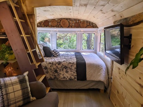 Cozy interior of a tiny home with a bed, wooden accents, and large windows overlooking greenery.