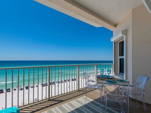 A balcony with a glass table and chairs overlooking a sandy beach and turquoise ocean under a clear blue sky.