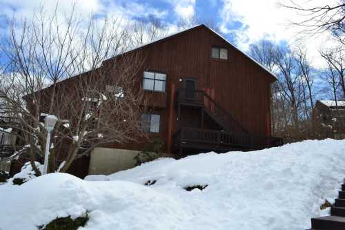 A wooden house on a snowy hillside, with a staircase leading to the entrance and trees in the background.