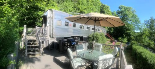 A vintage train car converted into a cozy outdoor space with a patio, table, and umbrella surrounded by greenery.
