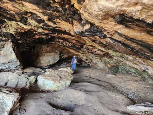 A person stands inside a large cave with rocky walls and a sandy floor, surrounded by natural formations.