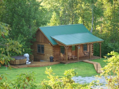 A cozy wooden cabin with a green metal roof, surrounded by trees and featuring a porch and hot tub.