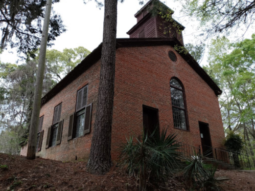 A historic brick building surrounded by trees, featuring a bell tower and large windows.