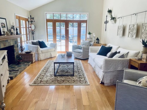 Bright living room with white sofas, a coffee table, and large windows overlooking a patio. Warm wooden floors and decor.