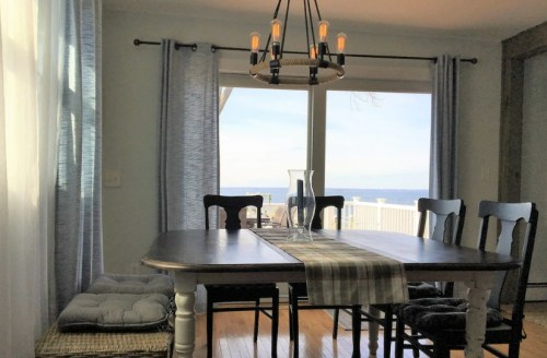 A dining area with a wooden table, chairs, and a chandelier, overlooking a scenic view of the water through large windows.