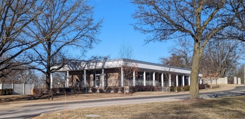 A modern building with a flat roof, surrounded by trees and a clear blue sky, set back from a winding road.