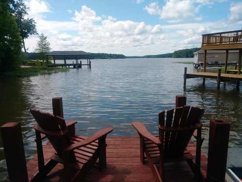 Two wooden chairs on a dock overlooking a calm lake with trees and clouds in the background.