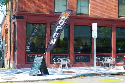 A restaurant with large windows and an "OPEN" flag outside, featuring outdoor seating and a chalkboard menu.