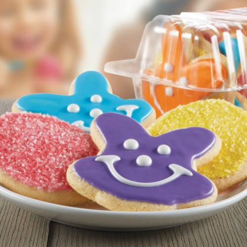 Colorful, decorated cookies in a plate, featuring smiley faces and vibrant icing, with a container of more treats in the background.