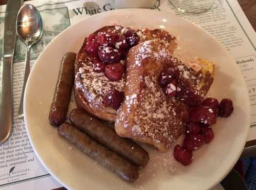 A plate of French toast topped with powdered sugar and cranberries, accompanied by two sausage links.