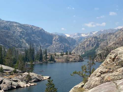 A serene lake surrounded by rocky mountains and pine trees under a clear blue sky. Snow-capped peaks in the distance.
