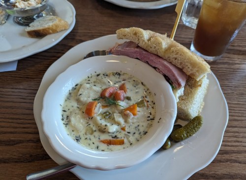A bowl of creamy soup with vegetables beside a sandwich filled with ham on a plate, accompanied by pickles.