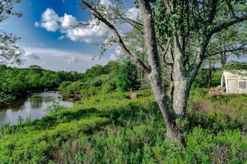A serene landscape featuring a river, lush greenery, and a tent under a blue sky with scattered clouds.
