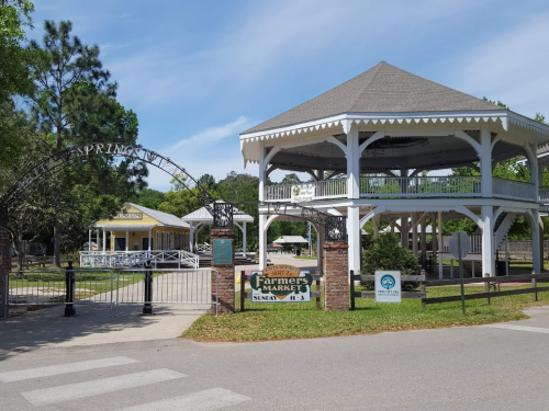A scenic view of a farmers market entrance with a gazebo and greenery under a clear blue sky.