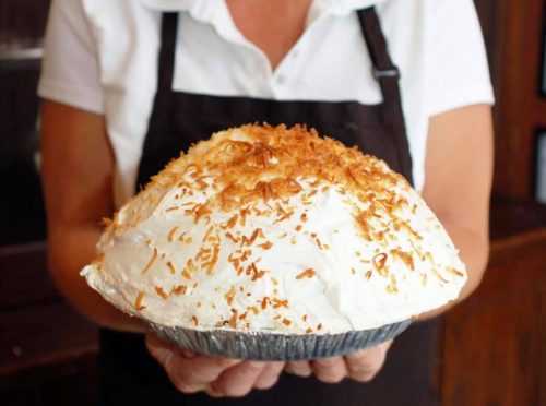 A person holds a large coconut cream pie topped with toasted coconut flakes.