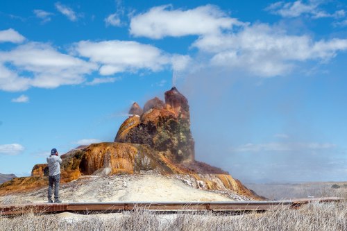 A person photographs a colorful geothermal feature with steam rising against a blue sky and scattered clouds.