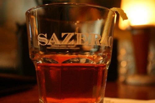 A close-up of a Sazerac cocktail in a glass, garnished with a lemon twist, on a wooden table.