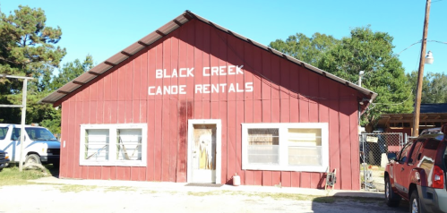A red wooden building with "Black Creek Canoe Rentals" sign, surrounded by trees and parked vehicles.