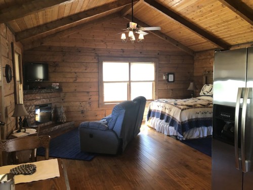 Cozy cabin interior with wooden walls, a bed, recliner, TV, and a large window letting in natural light.