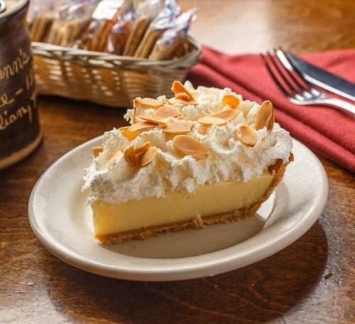 A slice of creamy pie topped with whipped cream and almond slices, served on a white plate with a basket of cookies in the background.