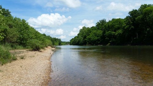 A serene river scene with a sandy shore, lush green trees, and fluffy clouds in a blue sky.