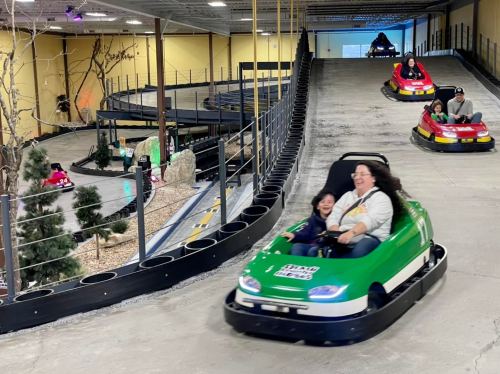 A woman and child enjoy a ride in a green go-kart on a winding indoor track, with other karts in the background.