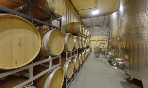 A wine cellar featuring stacked wooden barrels and stainless steel fermentation tanks in a spacious, well-lit room.