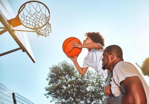 A child jumps to dunk a basketball while being lifted by an adult, with a clear sky and trees in the background.