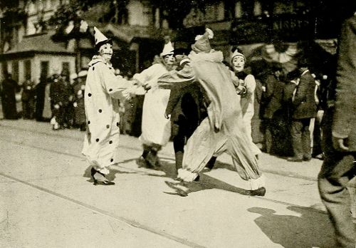 A vintage parade scene featuring performers in clown costumes dancing on the street, surrounded by onlookers.