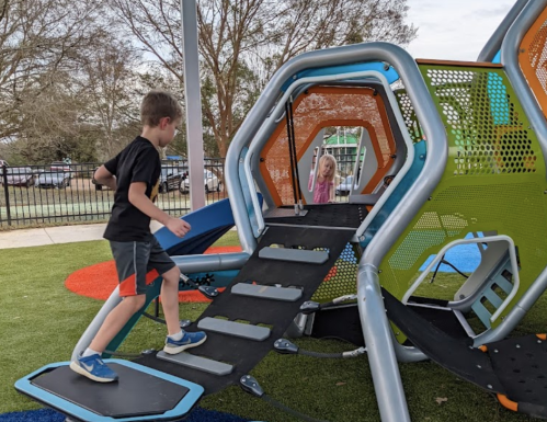 Two children play on a colorful playground structure, one climbing while the other watches from inside.