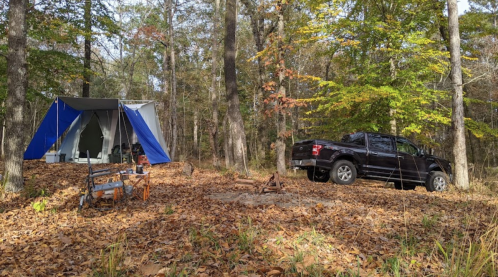 A campsite in the woods featuring a blue tent, a parked truck, and a fire pit surrounded by autumn leaves.