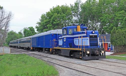 A blue train engine pulls a passenger car along a track, surrounded by greenery and trees.