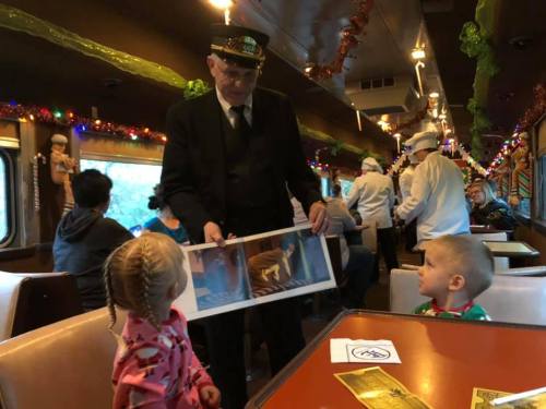 A train conductor shows a picture to two children in a festive train car decorated for the holidays.