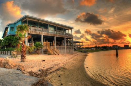 A beachside house at sunset, with palm trees and a sandy shore reflecting vibrant orange and yellow hues in the sky.