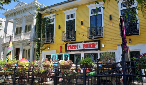 A vibrant yellow restaurant with a sign reading "Tacos & Beer," featuring outdoor seating and lush greenery.