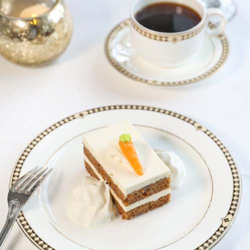 A slice of carrot cake with cream frosting and a small carrot decoration, served with whipped cream and a cup of coffee.