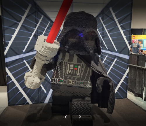 A large LEGO sculpture of a character in a black cape, holding a red lightsaber, set against a geometric background.
