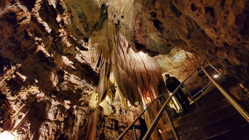 A person stands on stairs in a cave, surrounded by textured rock formations and soft lighting illuminating the walls.