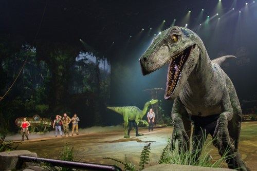 A dinosaur show featuring a large dinosaur and performers on stage in a jungle-themed setting.