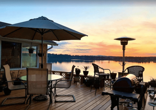 A serene lakeside deck at sunset, featuring a table, chairs, and a grill, with vibrant colors reflecting on the water.